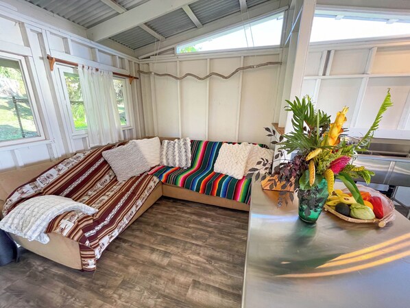 A cozy, well-lit room with a large, colorful sofa, a wooden table with a fruit basket, and a view of greenery outside.