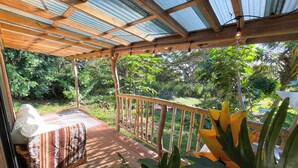 A covered outdoor deck with a wooden railing, a bench, and a table with a patterned tablecloth. - Kukui Cabin with Private Bath (Kealakekua)