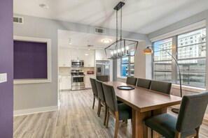 A modern kitchen with stainless steel appliances, a dining area with a wooden table and chairs, and large windows offering a view of buildings outside. - 4BD Oasis - Steps to French Quarter (New Orleans)