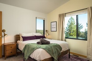 A bedroom with a bed, a nightstand, a window with a view of trees, and a framed quote on the wall.