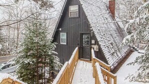 Un escalier en bois mène à un chalet aux couleurs sombres, doté d’une cheminée, et entouré d’arbres couverts de neige.