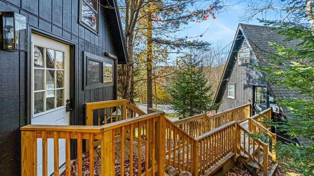 Une terrasse en bois mène à une maison aux couleurs sombres, dotée d’une porte et de fenêtres blanches.