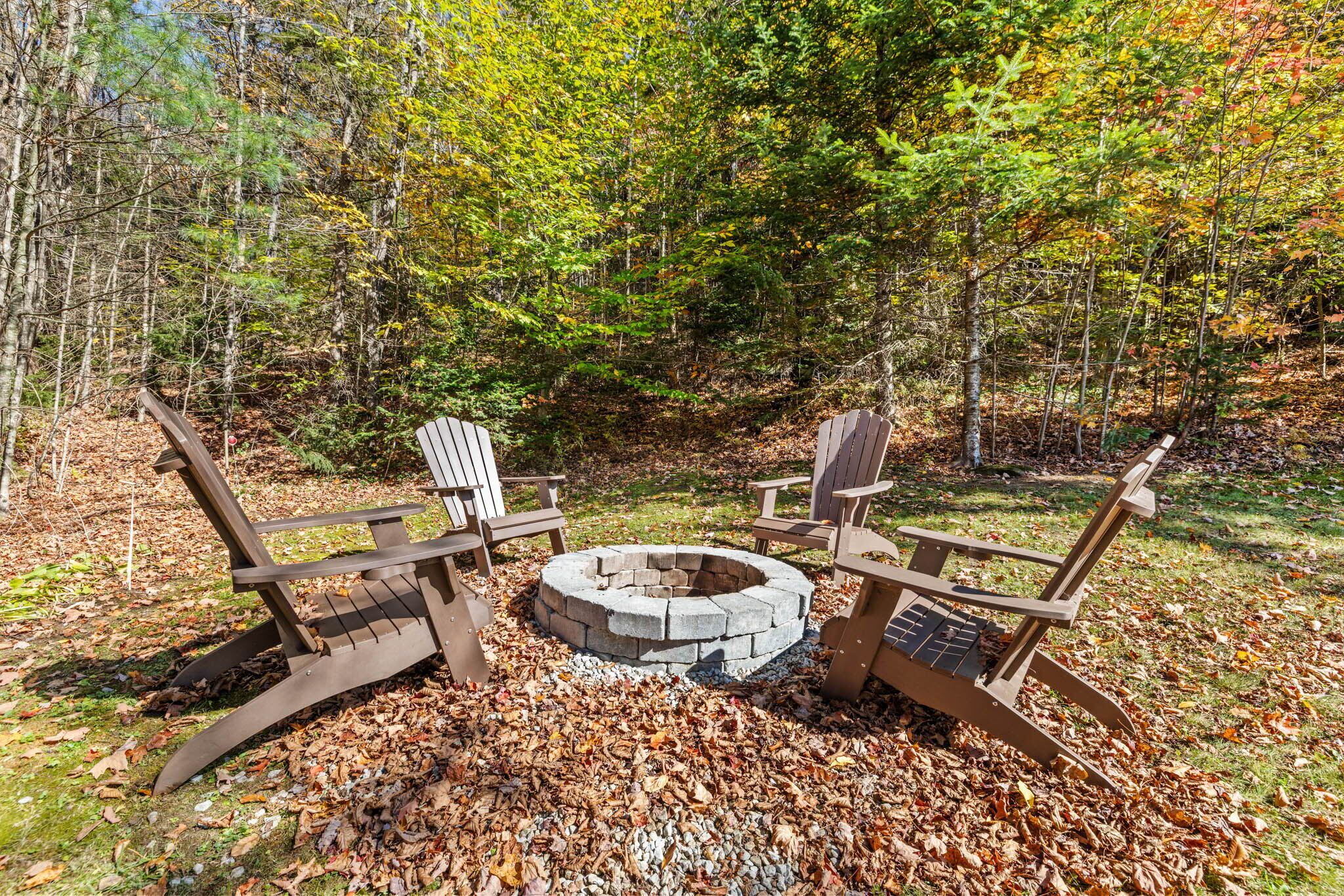A circular stone fire pit surrounded by Adirondack chairs in a forested area with fallen leaves.