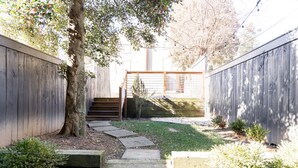 Un jardin à l’arrière de la maison, avec une clôture en bois, un arbre, un chemin de pierres et un petit espace aménagé en jardin.