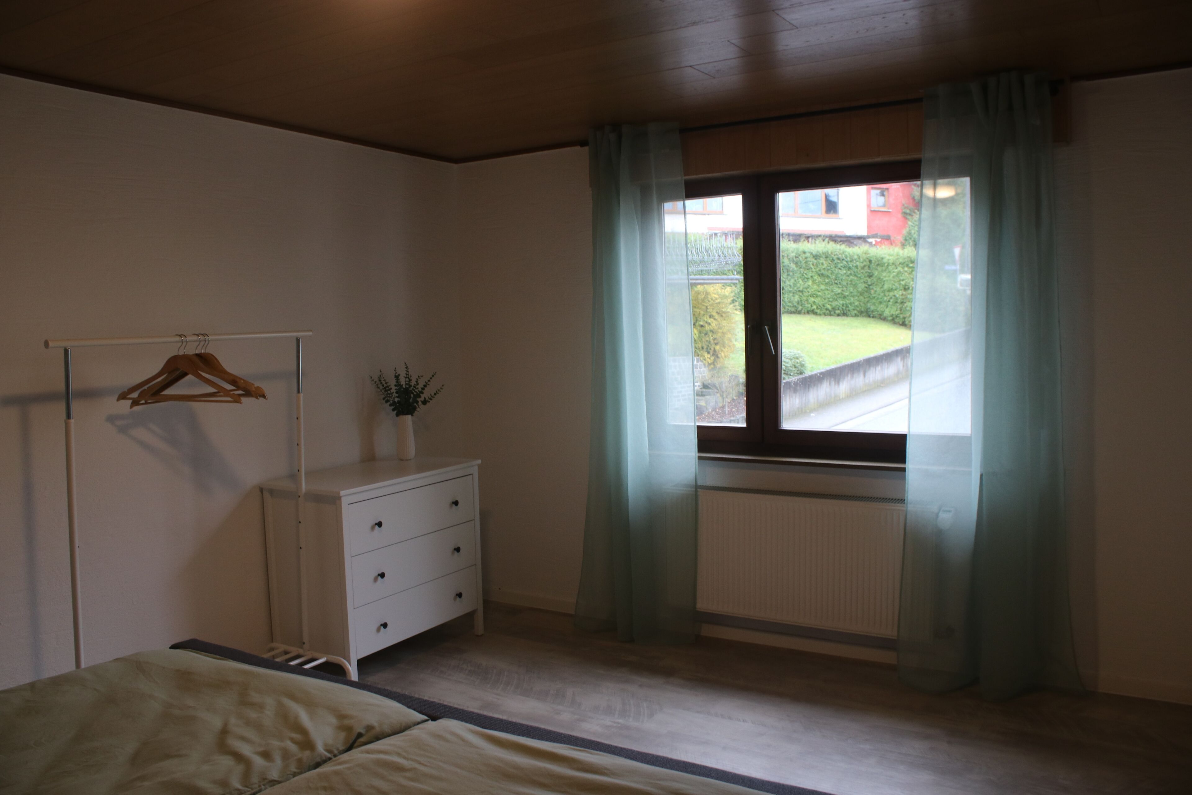 A bedroom with a bed, a dresser, a radiator, and a window with curtains.