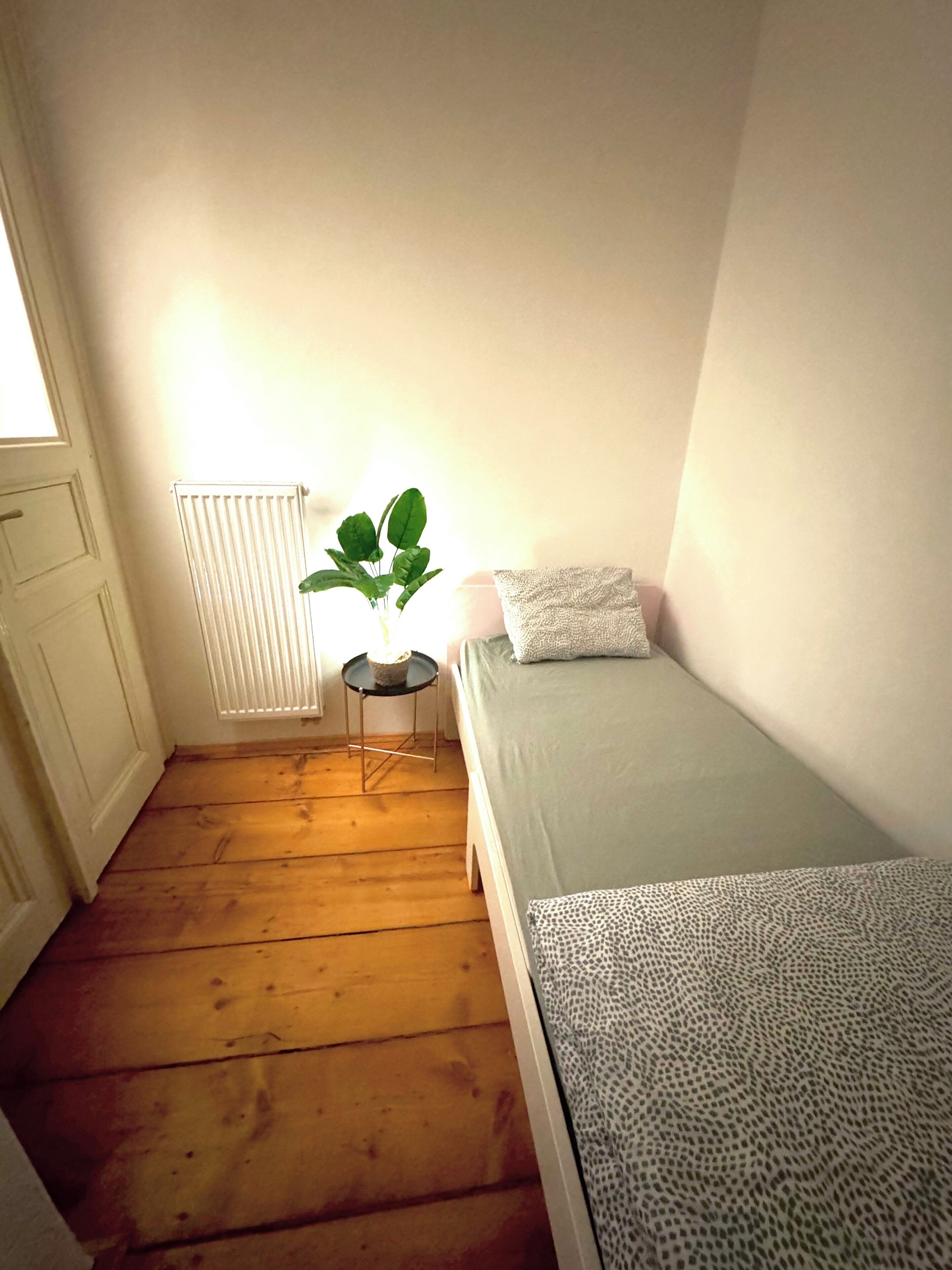 Une petite chambre avec un lit simple, un plancher en bois, un radiateur, une petite table avec une plante et une fenêtre.