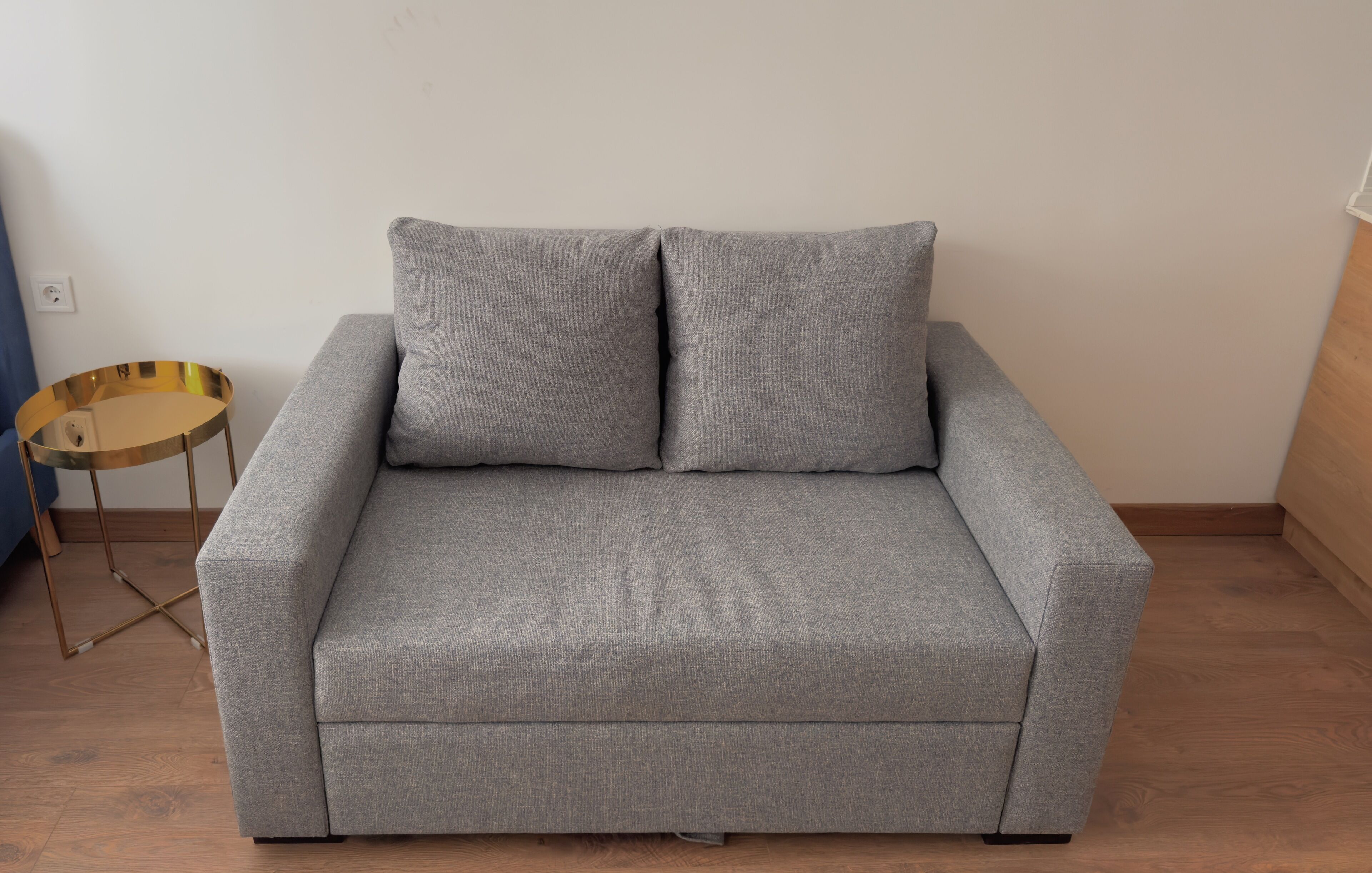 A grey sofa with two cushions in a room with wooden flooring and a small round table with a gold base.