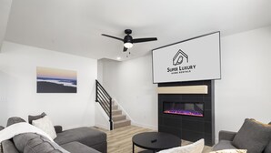 A modern living room with a fireplace, grey sofas, a staircase, and a ceiling fan. - New 7 bdrm home (sleeps 28) w/ lake views, sauna, hot tub, & outdoor dining! (HOLLISTER)