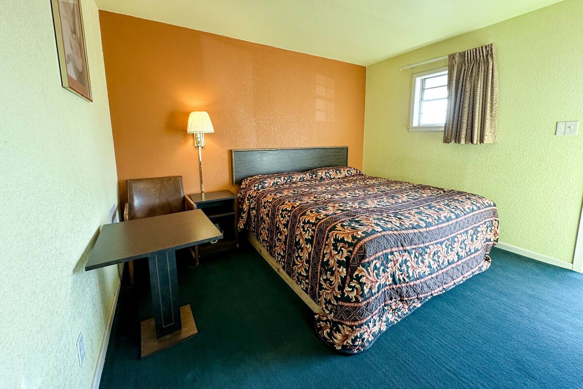 Ein Hotelzimmer mit Bett, Schreibtisch, Stuhl, Lampe und einem fenster mit Vorhängen.