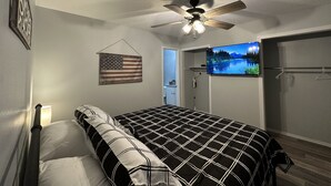 A bedroom with a bed, a ceiling fan, an American flag, a TV, and a door leading to a bathroom. - Piney Woods 6 - King Bed (Texarkana)