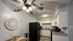 A compact kitchen with white cabinets, a black refrigerator, a wooden dining table with two chairs, and a ceiling fan. - Piney Woods 6 - King Bed (Texarkana)