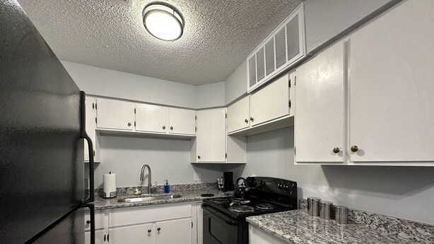 A kitchen with white cabinets, a black refrigerator, a stove, and granite countertops. - Piney Woods 6 - King Bed (Texarkana)
