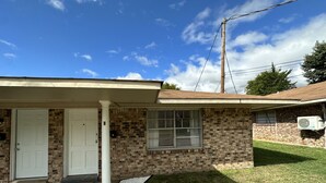 A single-story brick house with a front porch, a window with blinds, and a small front yard. - Piney Woods 6 - King Bed (Texarkana)