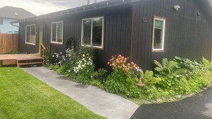 Ett hus med en mörk fasad, en träveranda och en välskött trädgård med olika sorters växter och blommor.