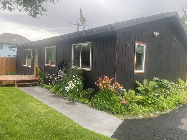 A house with a dark exterior, a wooden deck, and a well-maintained garden with various plants and flowers. -  The Modern Moose. A Ranch home close to airport. (Anchorage)