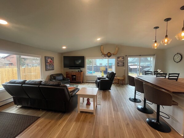A spacious living room with a large flat-screen TV, a wooden dining table with bar stools, a leather sofa, and a large window offering a view of the backyard. -  The Modern Moose. A Ranch home close to airport. (Anchorage)