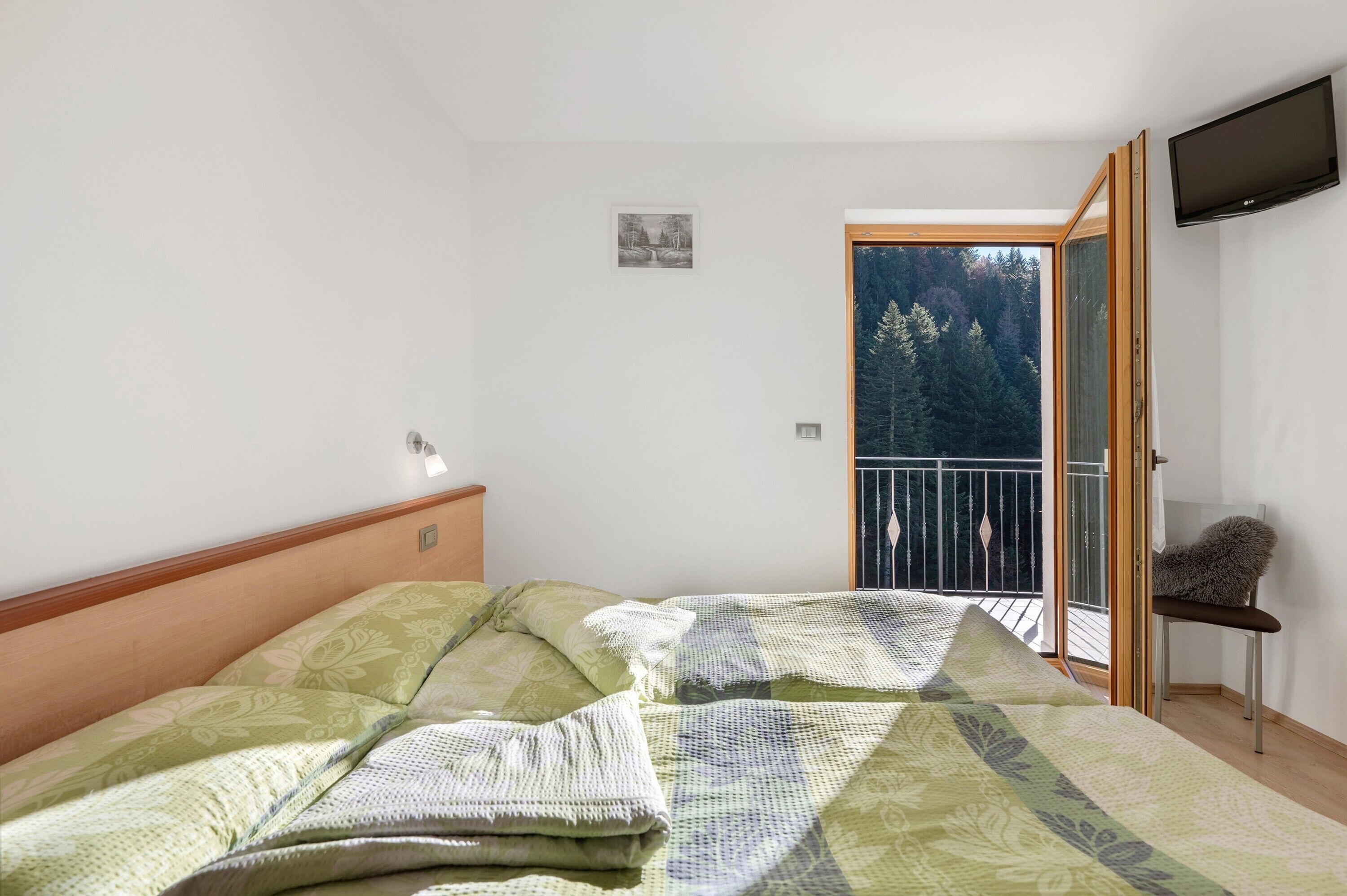 Een slaapkamer met een bed, een klein tafeltje, een stoel, een televisie en een balkon met uitzicht op bomen.