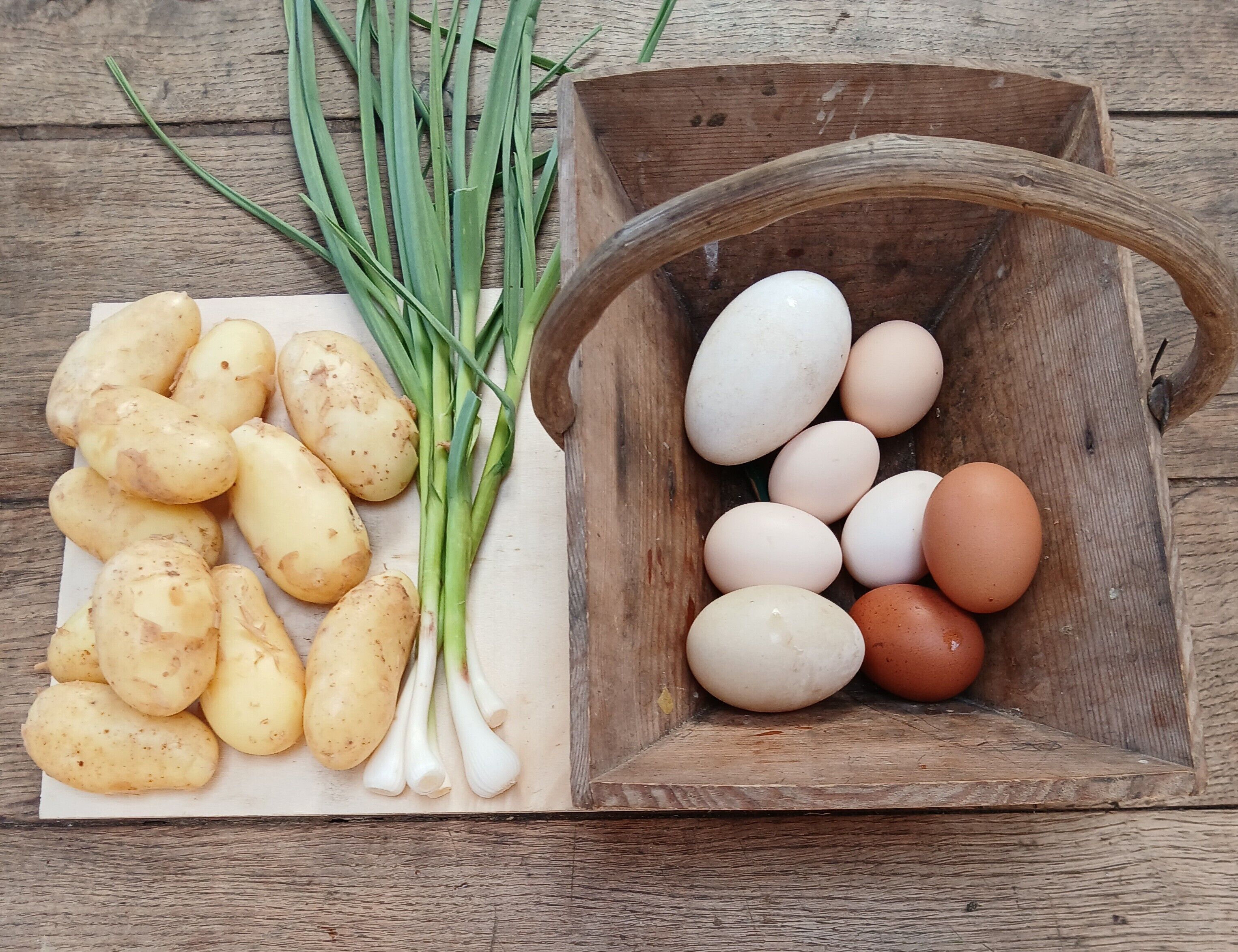 Une table en bois sur laquelle se trouve une planche à découper blanche avec des pommes de terre et des oignons verts, ainsi qu'un bol en bois contenant des œufs.