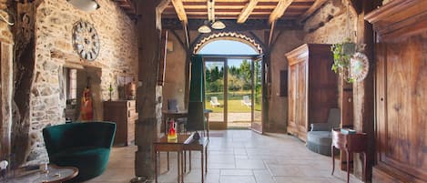 A rustic interior with stone walls, wooden beams, a dining table, and a view through an open archway.