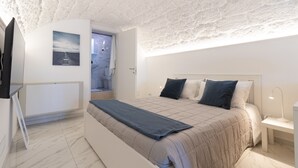 A bedroom with a bed, a bedside table, a nightstand, a wall-mounted TV, and a door leading to a bathroom.