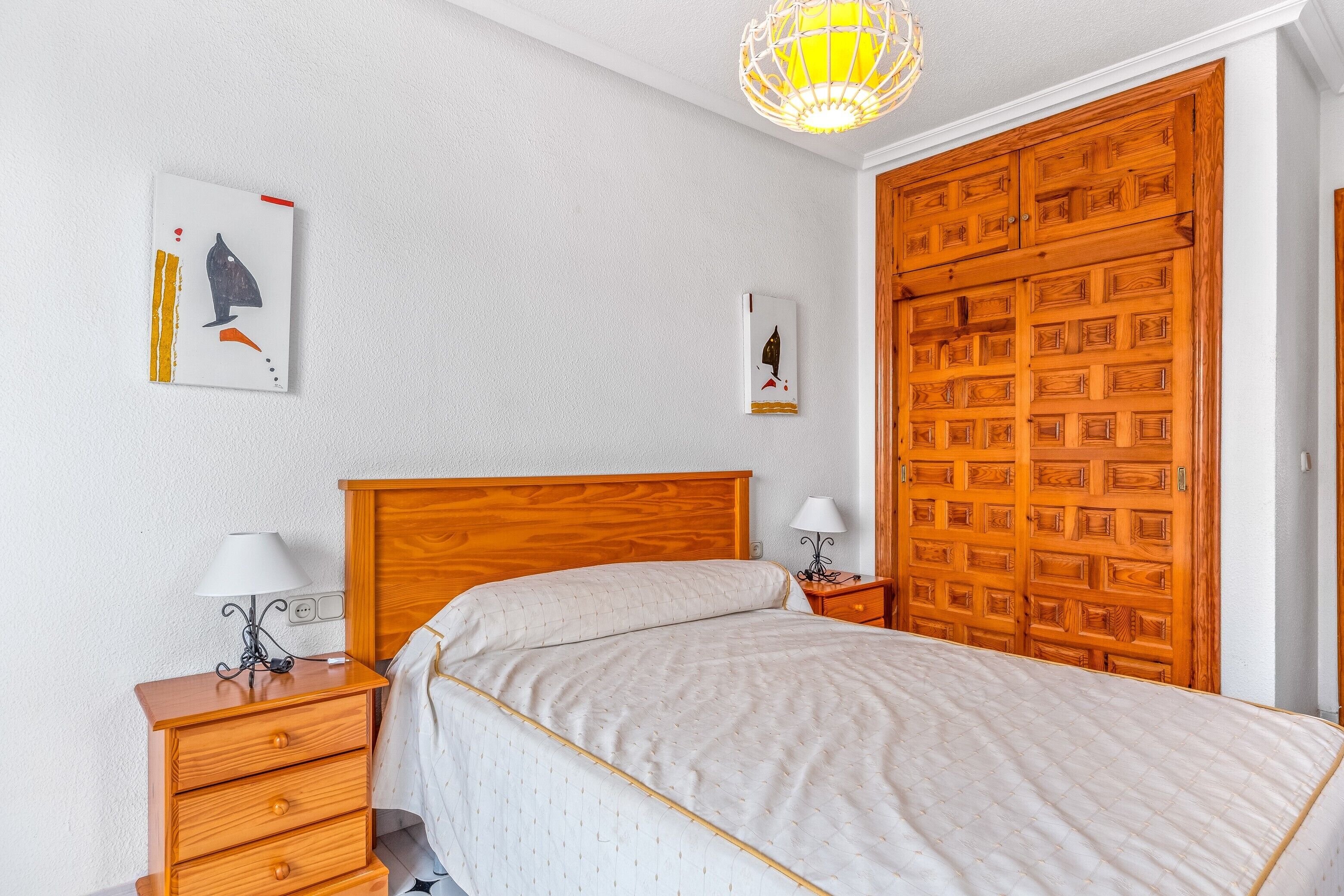 Un dormitorio con una cama de madera, dos mesitas de noche con lámparas, un armario de madera y dos obras de arte colgadas en la pared.
