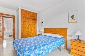 Une chambre avec une armoire en bois, un lit recouvert d’une couvre-lit à motifs floraux bleus, une table de chevet avec une lampe et une œuvre d’art accrochée au mur.
