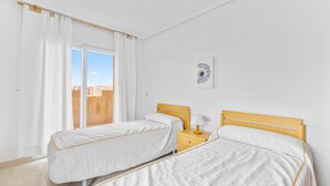 A room with two beds, a window with curtains, a bedside table with a lamp, and a wall-mounted artwork.