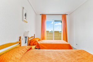 A room with two beds, a wooden headboard, a wall-mounted lamp, and a window with orange curtains.