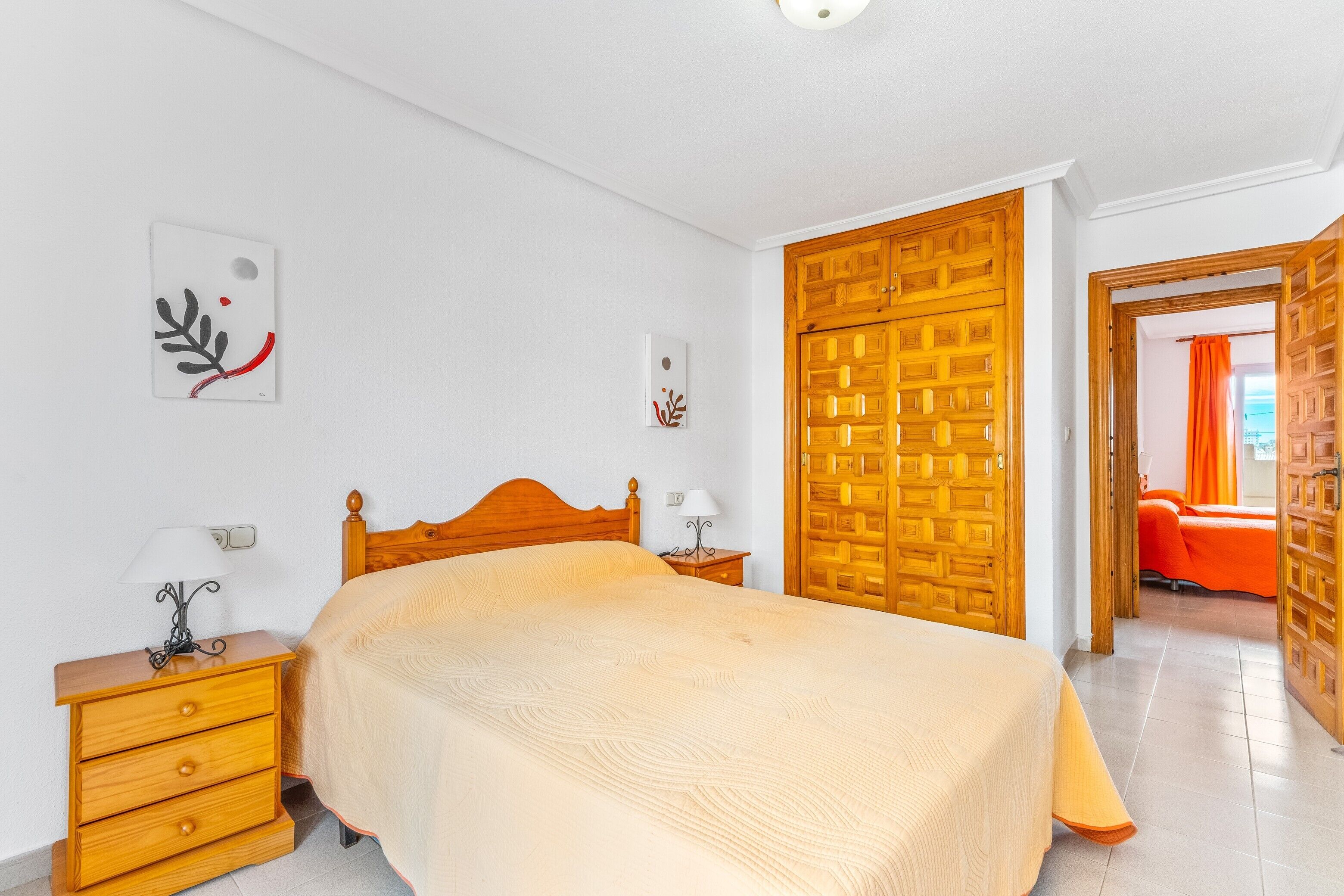 Une chambre à coucher avec un lit en bois, une table de chevet avec une lampe, une armoire en bois et une porte donnant sur une autre pièce.