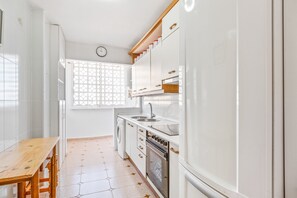 Une cuisine avec des meubles blancs, une table en bois, un lave-linge et une horloge au mur.