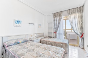 A bedroom with two beds, a nightstand, a lamp, a window with curtains, and a view of the outside.