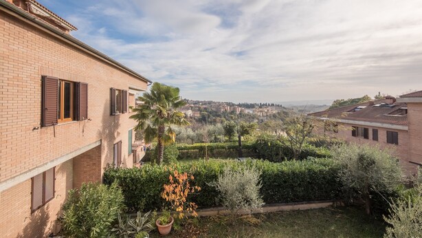 Un edificio residencial con fachada de ladrillo, múltiples ventanas con contraventanas y un jardín bien cuidado con diversas plantas y una palmera.