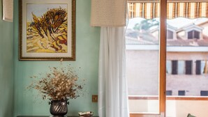 Una habitación con vistas a edificios, un cuadro en la pared, un florero con flores secas sobre una mesa y una silla.