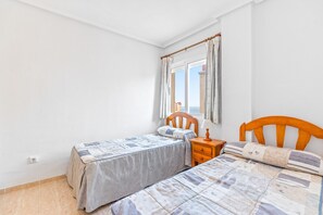 Une chambre avec deux lits simples, une tête de lit en bois, une table de chevet, une fenêtre avec des rideaux et un mur blanc.