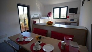 A compact room with a bunk bed, a kitchenette, a dining area with red placemats, and a window providing natural light.