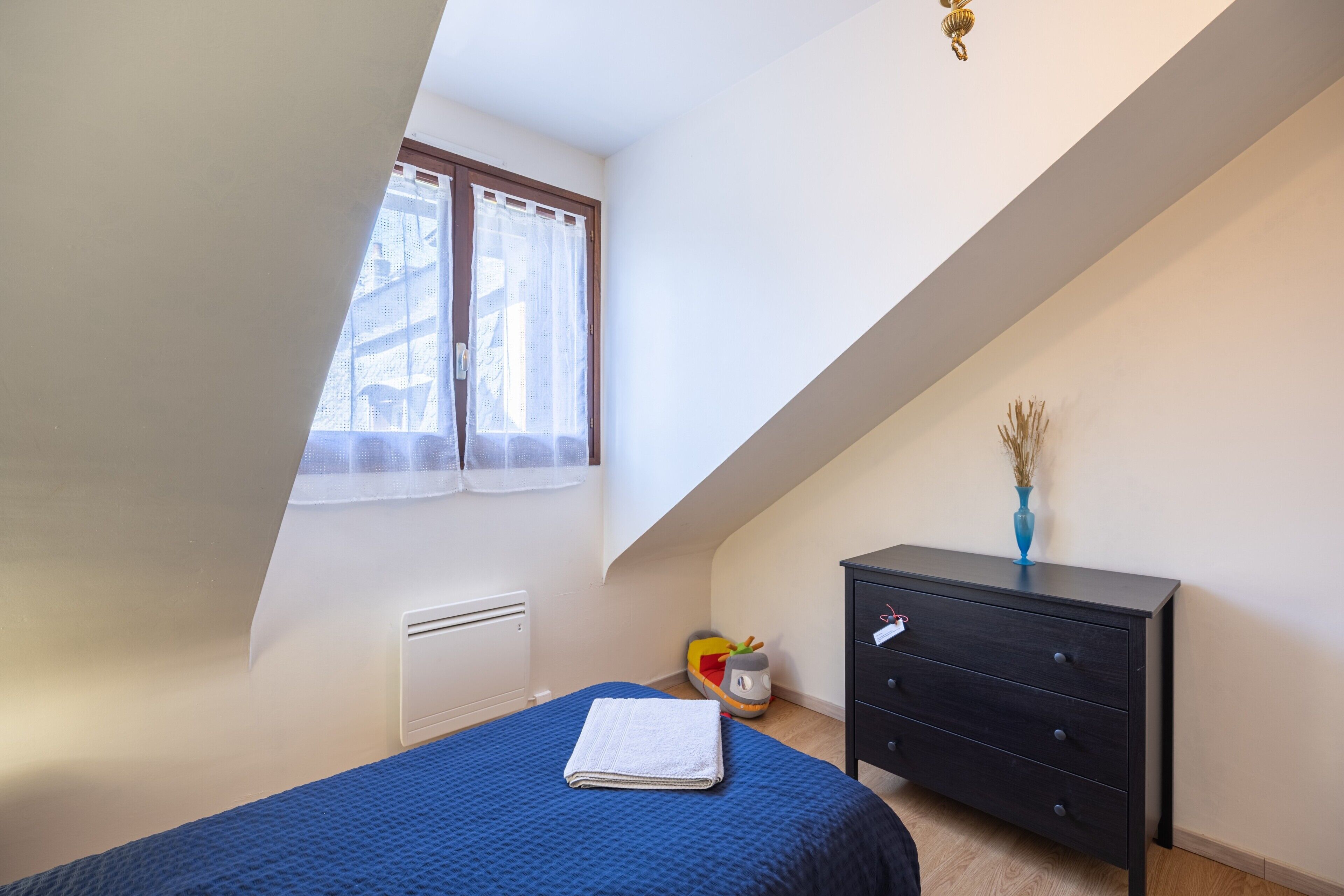 Un dormitorio con techo inclinado, una cama con cubrecolcha azul, un aparador, un radiador y una ventana con cortinas translúcidas.