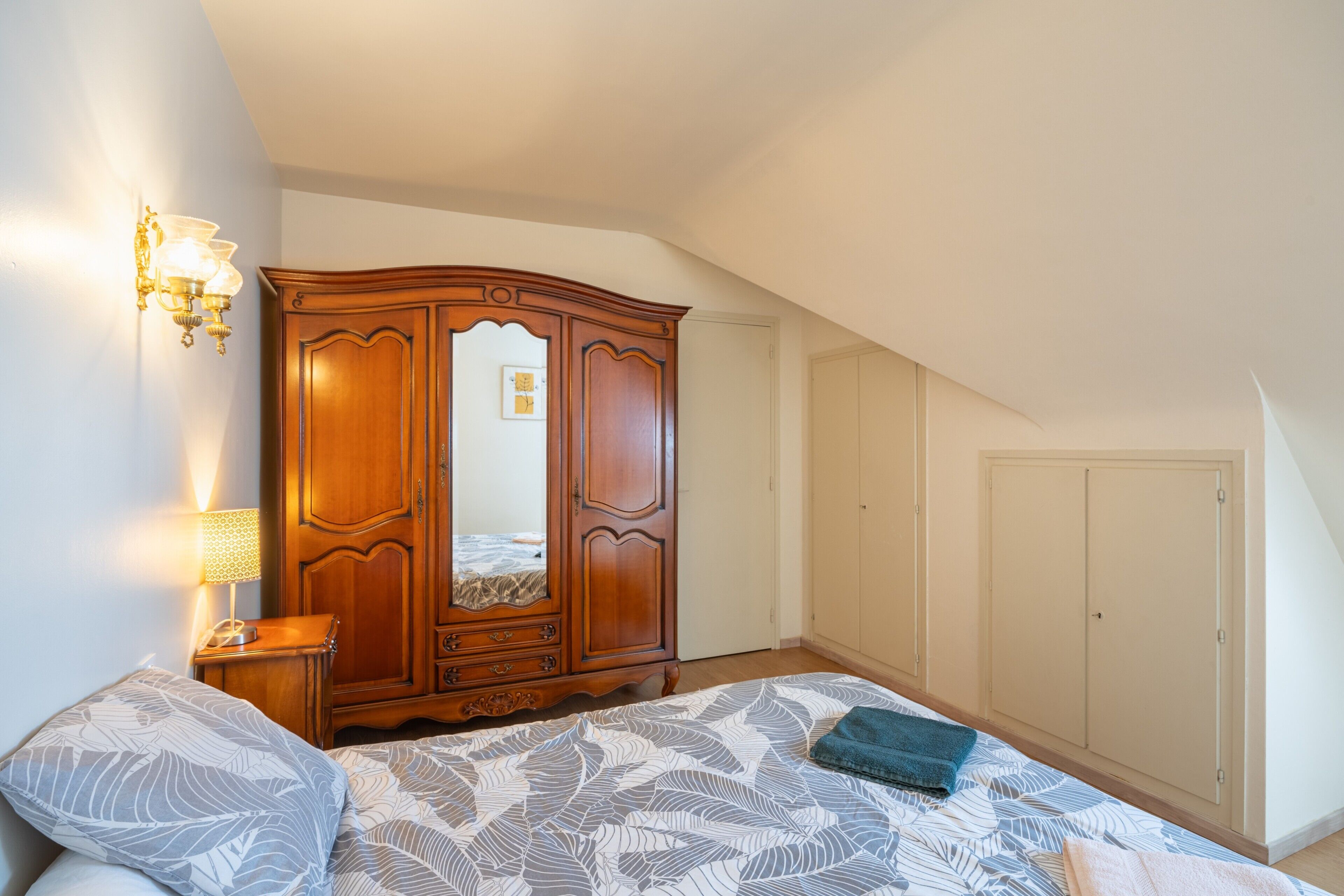 A bedroom with a bed, a wardrobe, a nightstand, and a wall-mounted light fixture.
