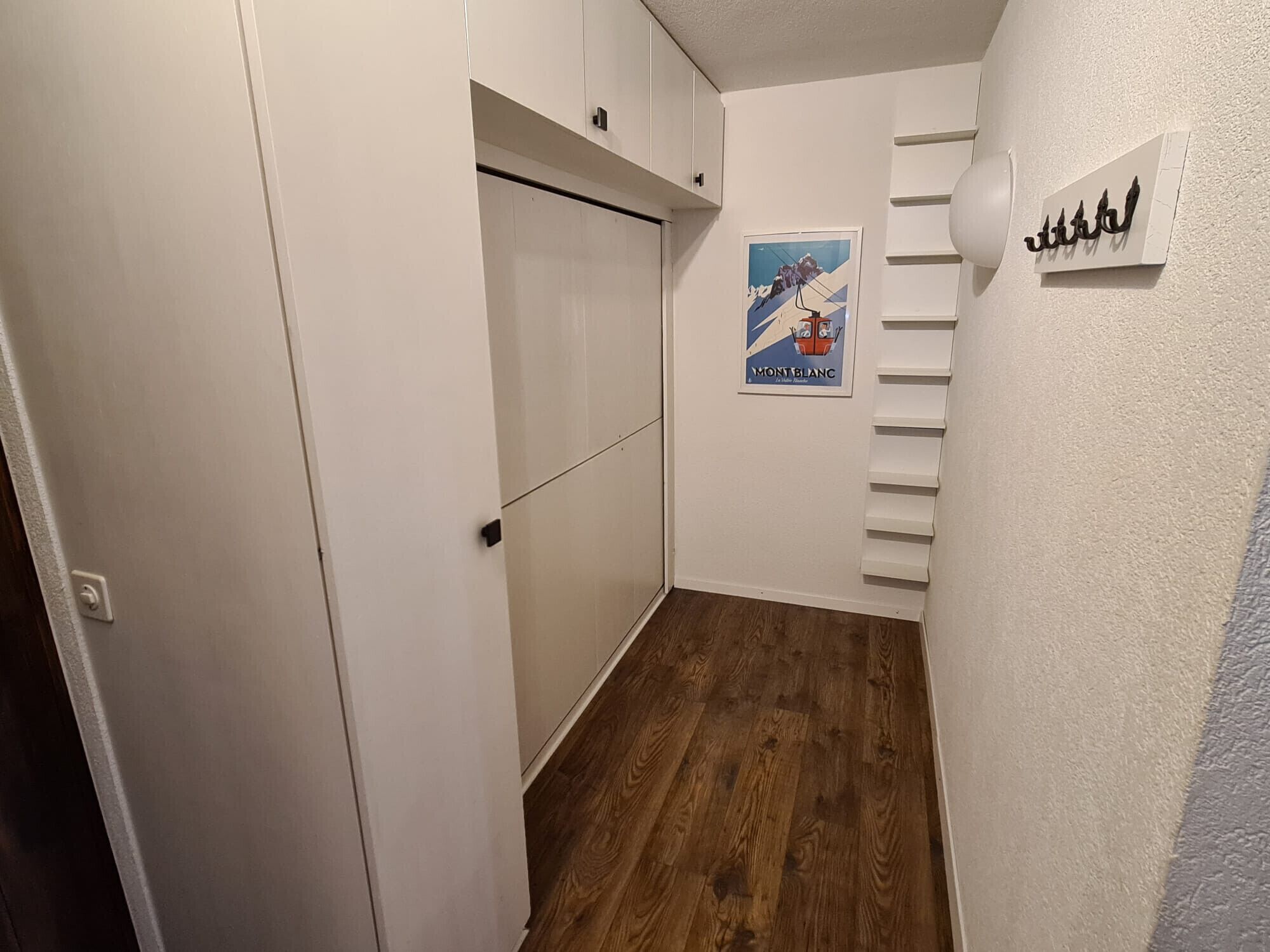 Un corridoio stretto con pavimento in legno, pareti bianche e una porta con maniglia nera.
