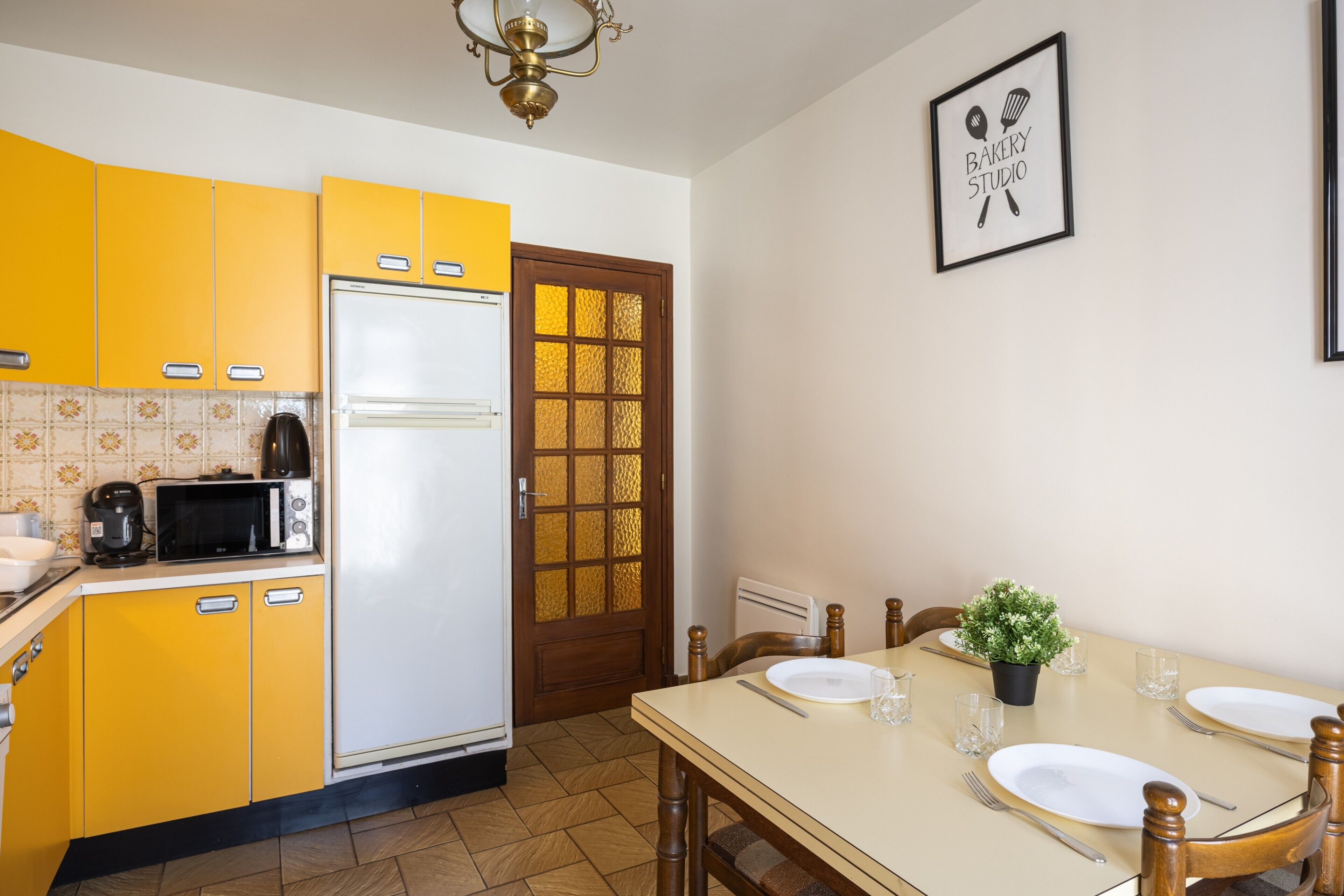 Une cuisine avec des meubles de couleur jaune, un réfrigérateur blanc, un four à micro-ondes et une table à manger dressée avec des assiettes et des couverts.