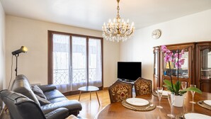 Ein Wohnzimmer mit einer Couch, einem Esstisch für vier Personen, einer Deckenleuchte, einem Fernseher und einem großen Fenster mit Vorhängen.