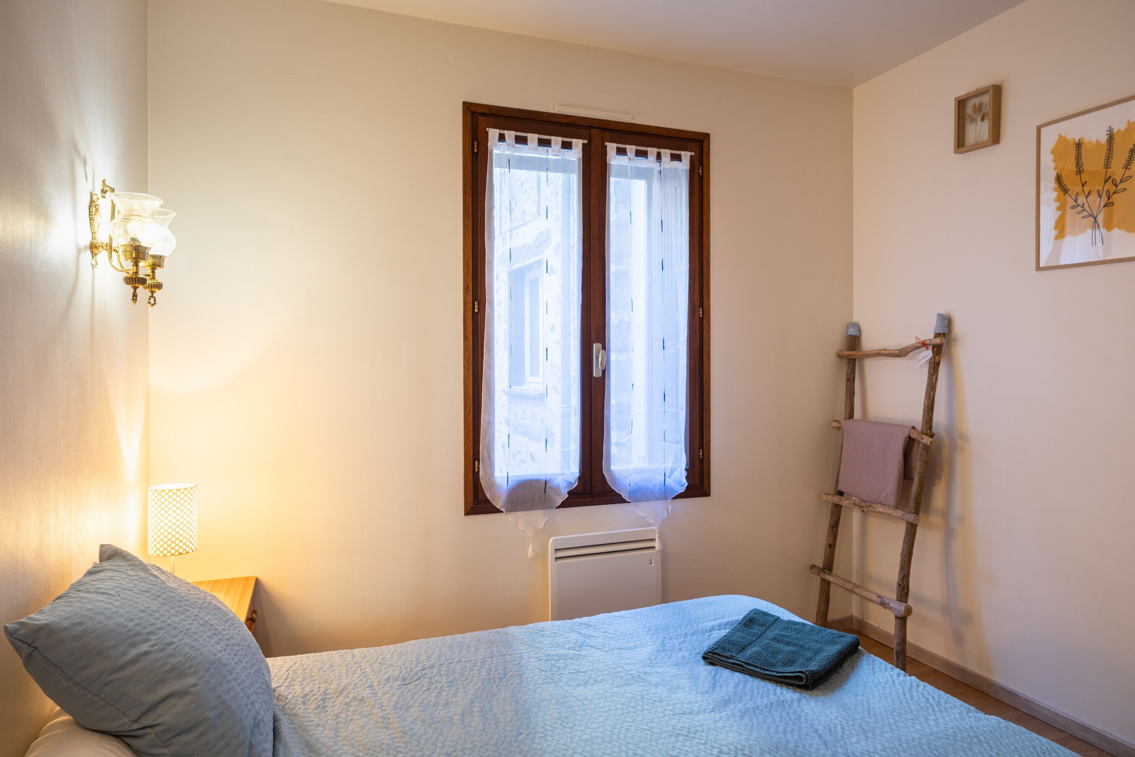 Um quarto com uma cama, um abajur, uma janela com cortinas, um radiador, uma escada de madeira e um quadro na parede.