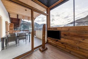 A modern interior with a wooden ceiling, a dining area with a glass table and chairs, a mounted TV, and a view of mountains through large windows.