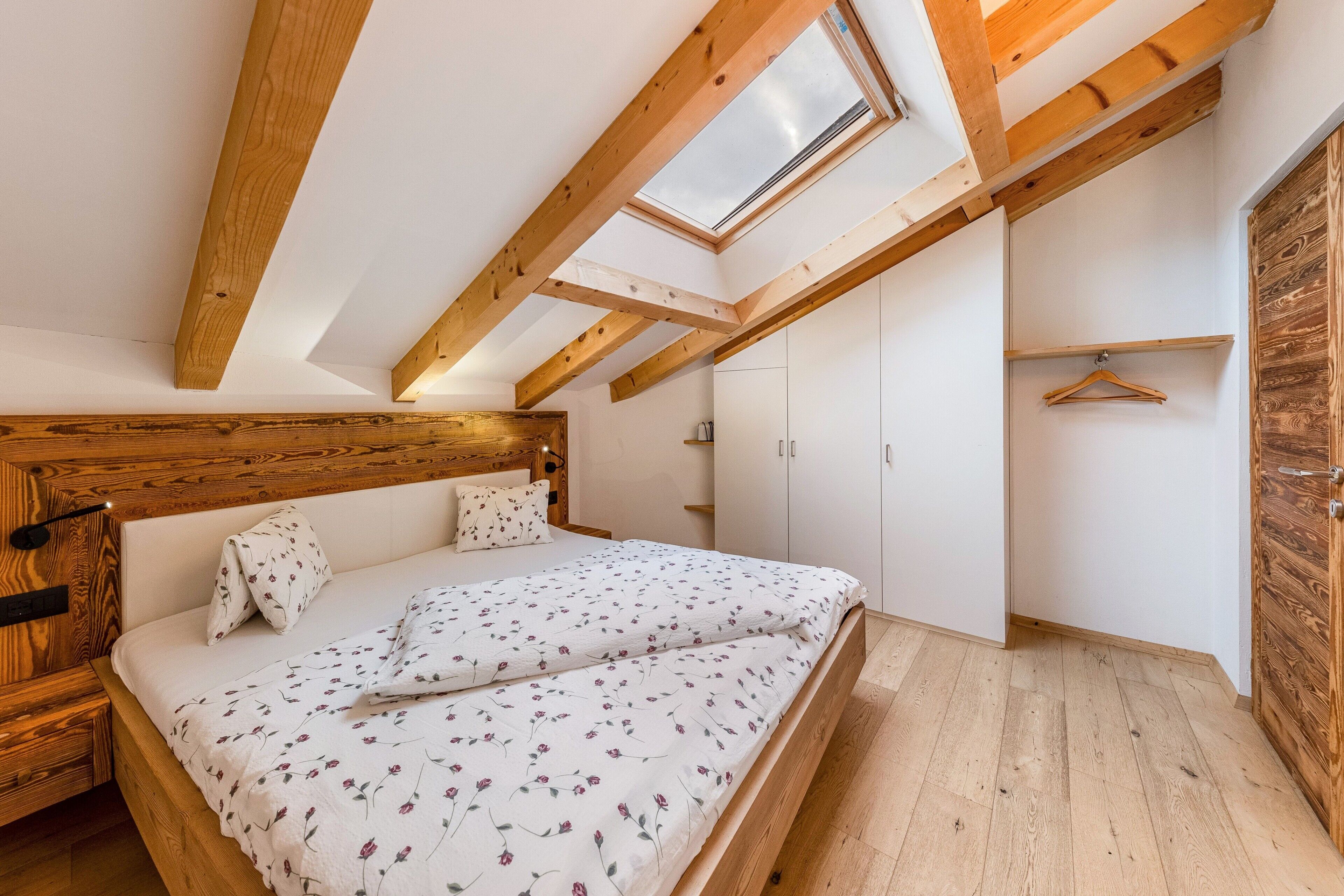 Une chambre avec un plafond en pente, des poutres en bois, un puits de lumière, un lit avec une literie à motifs floraux, une armoire et une porte en bois.