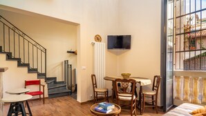 A living room with a staircase, a TV mounted on the wall, a dining table with chairs, and a window with a view.