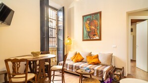 A cozy living room with a sofa, a dining table with chairs, a television mounted on the wall, and a painting above the sofa.