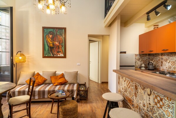 Un salon comprenant un canapé, des fauteuils, une petite table et un coin cuisine avec un mur recouvert de carreaux.
