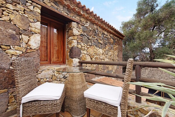 A stone building with a wooden door, a balcony with wicker furniture, and a view of trees.