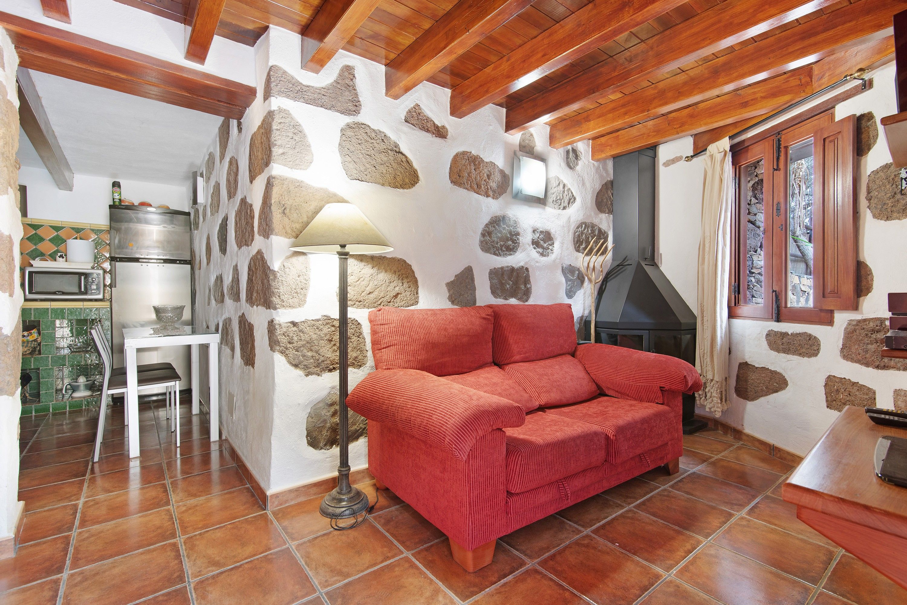 Una sala acogedora con pared de piedra, un sofá rojo, una mesa con lámpara y una cocina equipada.