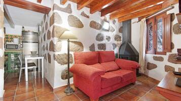 Una sala acogedora con pared de piedra, un sofá rojo, una mesa con lámpara y una cocina equipada.