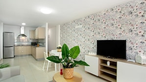 Une cuisine moderne dotée d’une cuisinière intégrée, un coin repas avec une table et des chaises blanches, un salon avec un téléviseur et un meuble de divertissement, et une grande plante en pot.